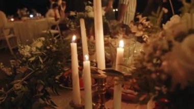 Close-up of a laid table with burning candles and bouquets of flowers at a party with many guests. Slow motion