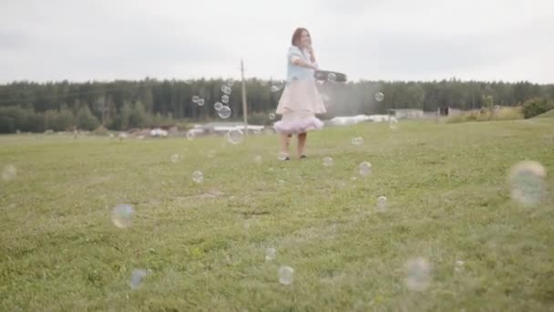 Happy beautiful young girl blows bubbles through a large circle in a meadow surrounded by a forest and a little girl catches them — Vídeo de Stock