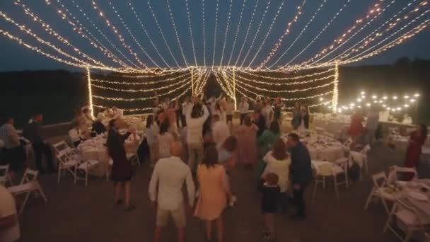 Belarus. MinskAugust 21, 2021: Guests have fun and dance in an outdoor area decorated with garlands with burning lights during the reception after the wedding ceremony — Video Stock