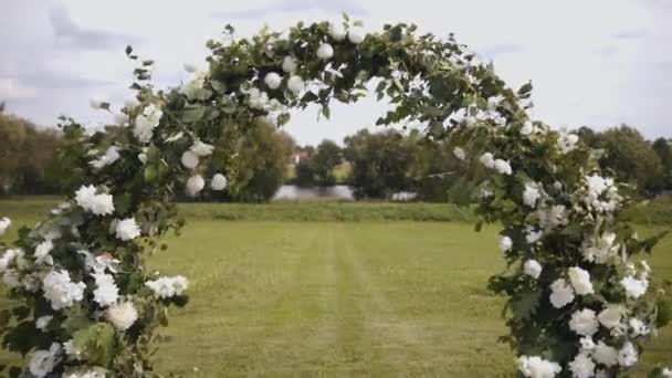 Primer plano de la parte superior del arco decorado con flores blancas y ramas de vegetación en el césped junto al río para una ceremonia de boda — Vídeos de Stock