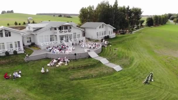 Вид сверху на террасу красивого усадебного дома с гостями и женихом с невестой за накрытыми столами на свадьбе после церемонии — стоковое видео