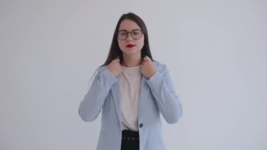 Siyah gözlüklü, kendine güvenen genç bir iş kadını beyaz arka planda duruyor ve kıyafetlerini ve saç stilini düzeltiyor.