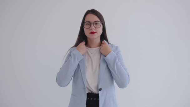 En självsäker ung affärskvinna i svarta glasögon står på en vit bakgrund och rätar ut sina kläder och frisyr — Stockvideo