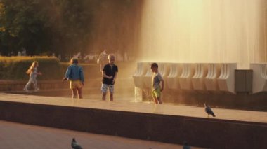 Minsk, Belarus - 29 Haziran 2021: Şehir meydanında gün batımında fıskiyenin damlaları ve su kenarındaki korkulukta koşan çocuklar. Yavaş çekim