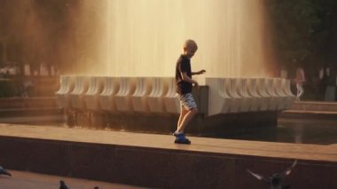 Minsk, Belarus - 29 Haziran 2021: Çocuklu aileler gün batımında şehir parkındaki çeşmenin yanında yürürken anaokulu öğrencisi bir çocuk güvercin kovalıyor. Yavaş çekim