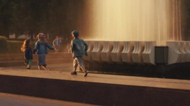 Minsk, Belarus - 29 Haziran 2021: Çocuklar gün batımında şehir parkındaki çeşmenin etrafındaki mermer korkulukta yürüyüp su damlalarını yakalarlar. Yavaş çekim