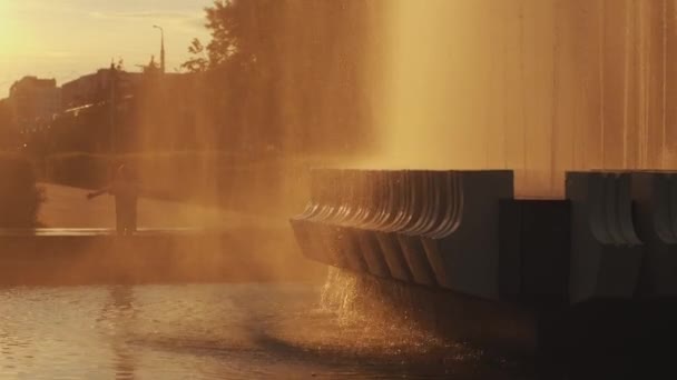 Un niño está de pie con los brazos extendidos bajo gotas de agua de una fuente al atardecer en un parque de la ciudad. Movimiento lento — Vídeos de Stock