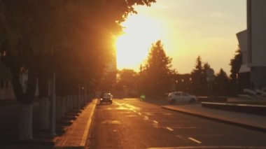Gün batımında yol boyunca sıra sıra ağaçlarla dolu bir şehir bloğunda arabalı bir yol. Ağır çekim. Kamera odağı değiştirir.