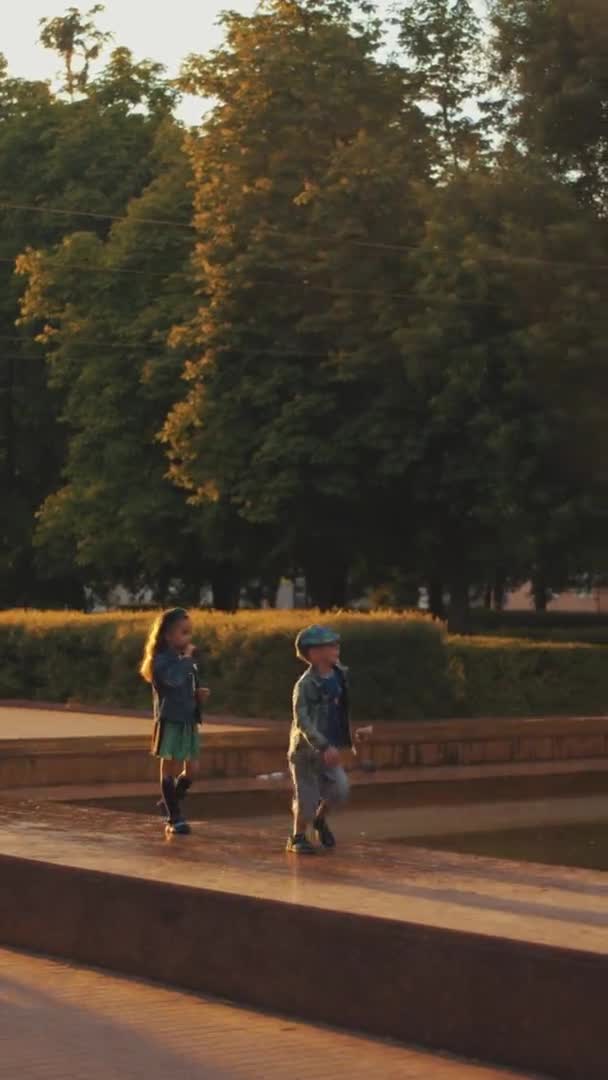 ベラルーシのミンスク- 2021年6月29日:子供たちは噴水の周りを走り、腕を伸ばして水の滴をキャッチします。垂直ビデオ。スローモーション — ストック動画