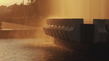 Bir çocuk gün batımında şehir parkında bir çeşmeden dökülen su damlalarının altında kollarını açmış duruyor. Yavaş çekim