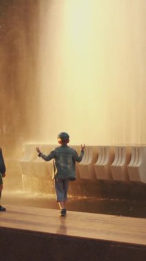 Minsk, Belarus - 29 Haziran 2021 anaokulu çağındaki neşeli kız ve oğlan gün batımında şelalenin korkuluğuna atlarlar. Dikey video. Yavaş çekim