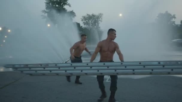 Két fiatal testépítő póló nélkül edz a vízsugár hátterében, és átugrik a tűzlépcsőn és fekvőtámaszokon. — Stock videók