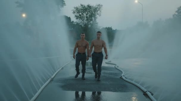Due uomini muscolosi con goccioline d'acqua sui loro busti nudi vanno tra getto d'acqua da tubi spray. Rallentatore — Video Stock