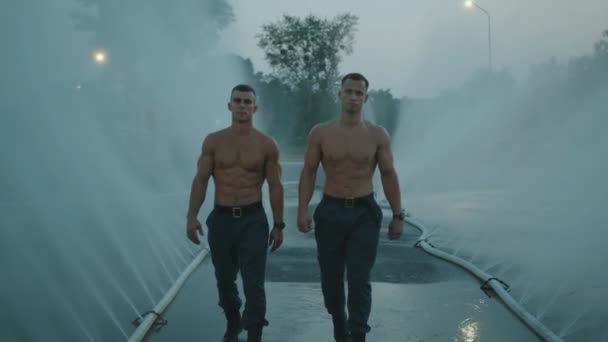 Two muscular men with water droplets on their naked torsos goes among water jet from spray hoses. Slow motion — Stock Video