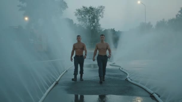 Two muscular firefighters with naked torsos run amid splashing water from spray hoses lying on the asphalt. Slow motion — Stock Video