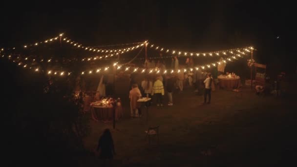 Luchtfoto van de pasgetrouwden dansen tussen de gasten op het gazon met gedekte tafels en rijen bloemenslingers met brandende gloeilampen — Stockvideo