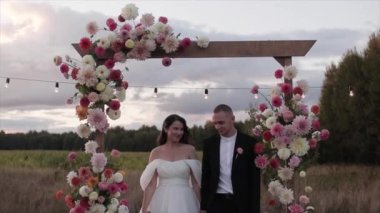 Mutlu gelin ve damat çiçek kemerinden el ele tutuşup doğadaki bir düğün töreninde birbirlerine bakarlar. Yavaş çekim