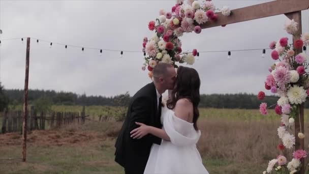 Küsschen des glücklichen Brautpaares am Blumenbogen mit Dahlien und Feuerwerk mit buntem Konfetti. Zeitlupe — Stockvideo