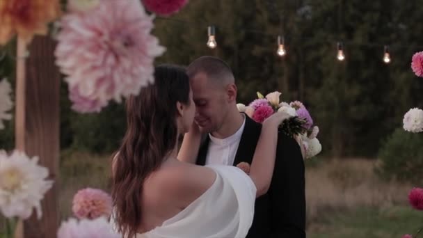 Un encantador retrato de los recién casados que se abrazan y el novio besa a la novia en el arco con dalias. Movimiento lento — Vídeo de stock