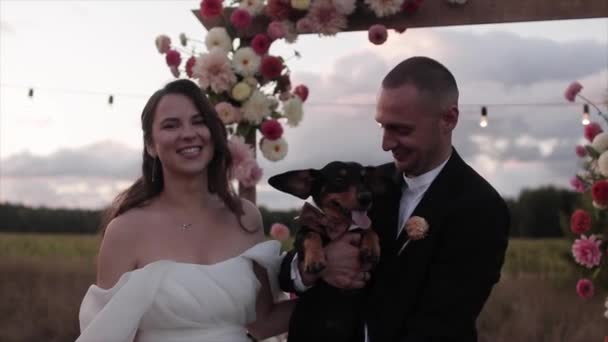 Roligt porträtt av skrattande nygifta med en tax i famnen nära en blomsterbåge på landet — Stockvideo