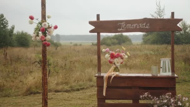 Ξύλινος πάγκος με δείκτη για λεμονάδα σε ένα γαμήλιο πάρτι στη φύση διακοσμημένα ανθοδέσμες από λουλούδια και ένα φανάρι — Αρχείο Βίντεο