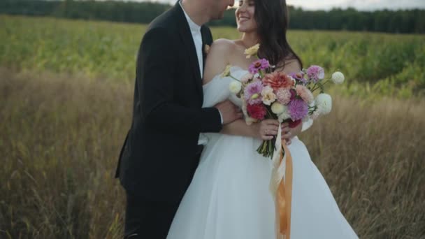 Ο γαμπρός φιλάει τη νύφη με ένα κομψό φόρεμα με ένα ντελικάτο μπουκέτο ντάλιες στην άκρη ενός καλαμποκιού — Αρχείο Βίντεο