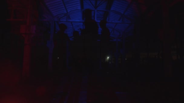 Grupa ludzi w ciemnym hangarze z metalowymi konstrukcjami w jasnoczerwonych i niebieskich reflektorach — Wideo stockowe