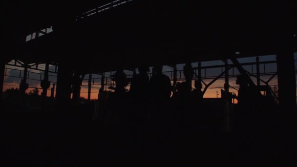 Silhuetas de um grupo de pessoas num grande edifício industrial abandonado com estruturas metálicas ao pôr-do-sol — Vídeo de Stock