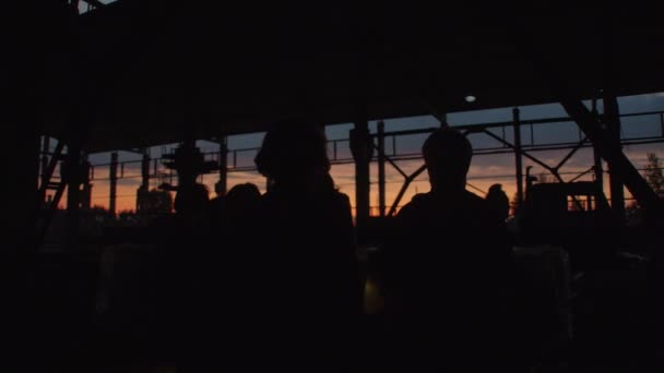 Czarne sylwetki grupy ludzi o zachodzie słońca w opuszczonym budynku magazynowym o konstrukcji metalowej — Wideo stockowe