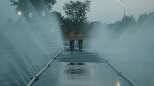 Çıplak gövdeli iki kaslı itfaiyeci asfaltta yatan sprey hortumlardan akan suyun arasında koşuyor. Yavaş çekim — Stok video