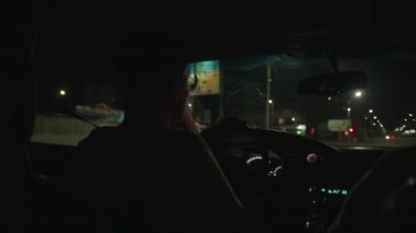 Araba kullanan gözlüklü genç bir adam ve arabanın ön camından yaz gecesinin manzarası.