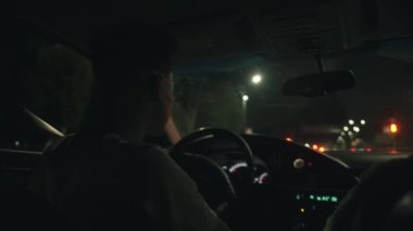 Gösterge paneli ışıkları ve gözlüklü sürücü içeriden ve şehir yolundan fener ve trafik ışıklarıyla görülüyor