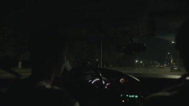 Gözlüklü genç bir adam geceleyin şehirde bir araba sürüyor. Otomobilin içinden ışıkların ve yoğun trafiğin görüntüsü