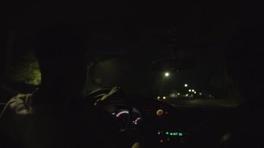 Gözlüklü genç bir adam geceleyin şehirde bir araba sürüyor. Otomobilin içinden ışıkların ve yoğun trafiğin görüntüsü