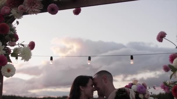 La mariée avec un bouquet dans ses mains étreint le marié et il l'embrasse sur le front dans l'ouverture de l'arche décorée de dahlias — Video