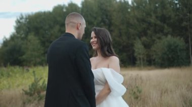 Mutlu gelin ve damat el ele tutuşup bir tarlanın ve ormanın arka planına karşı birbirlerinin gözlerine bakarlar.