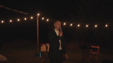 Damat, düğün töreninde açık alanda yanan ampullerle çelenklerin altında bir mikrofona konuşur.