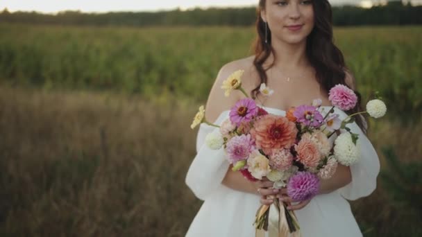 Uma linda noiva sorridente fica perto do campo e traz um delicado buquê de dálias com fitas para o rosto — Vídeo de Stock