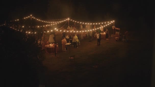 Luchtfoto van de pasgetrouwden dansen tussen de gasten op het gazon met gedekte tafels en rijen bloemenslingers met brandende gloeilampen — Stockvideo