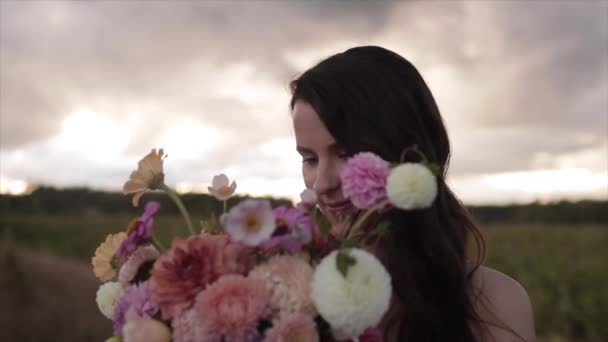 Portrét půvabné šťastné nevěsty s velkou kyticí dahlias u její tváře na rozmazaném pozadí přírody. Zpomalený pohyb — Stock video