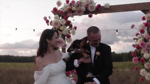 Funny portrait of laughing newlyweds with a dachshund in their arms near a flower arch in the countrysidere — Stock Video