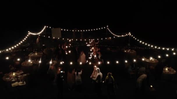 Minsk, Wit-Rusland - 29 juni 2021: Luchtfoto van pasgetrouwden die dansen omringd door gasten met sterretjes in een open ruimte versierd met brandende bloemenslingers en kaarsen — Stockvideo