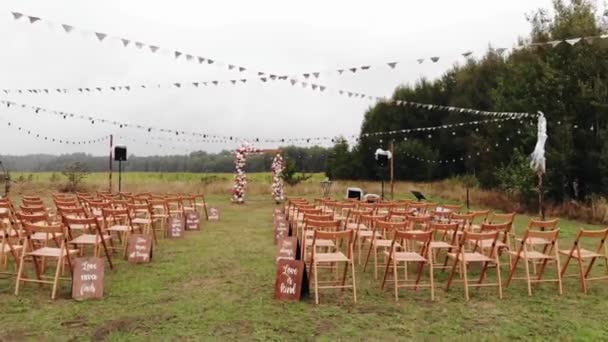 Imágenes de drones de filas de sillas y un arco de flores de madera en un lugar de ceremonia de boda en un césped cerca de un bosque — Vídeos de Stock