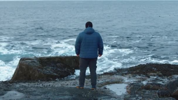 En man står på en stenig strand och havsvågorna bryter mot klipporna. Bakåt. Begreppet ensamhet och reflektion. Långsamma rörelser — Stockvideo