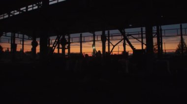 Gün batımında terk edilmiş bir depoda metal yapıları olan bir grup insanın siyah siluetleri.