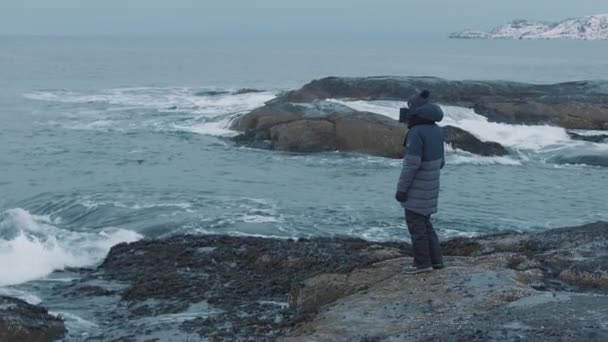 Ένας νεαρός άνδρας στέκεται σε μια βραχώδη ακτή και κοιτάζει τα κύματα που συντρίβονται στις πέτρες. Αργή κίνηση — Αρχείο Βίντεο