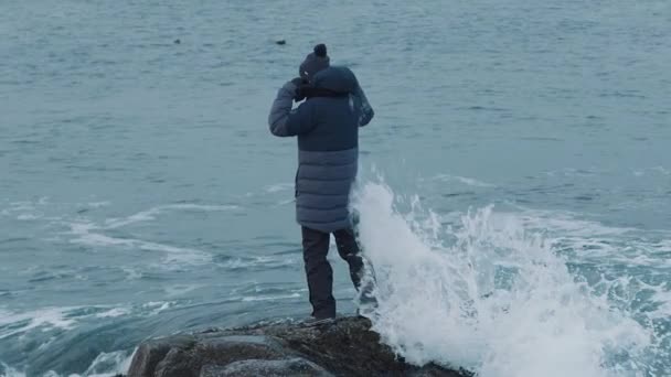 Un joven admira el paisaje marino y las olas que se rompen en las rocas inundan sus piernas. Movimiento lento — Vídeo de stock