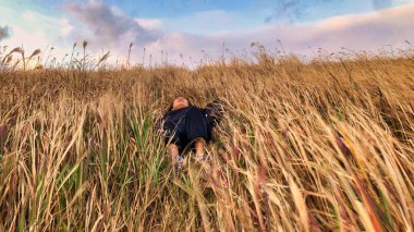 Asyalı bir kız bulutlu bir gökyüzünün rüzgarında buğday dağında uzanmış, yüzünde bir gülümsemeyle günbatımının tadını çıkarıyor.