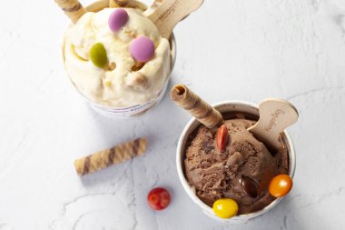 KYIV, UKRAINE - 7 Şubat 2022 Sundae ve Haagen-Dazs çikolatalı dondurmaları tahta çubuklarla kağıt kovalarda. Renkli süslemeler ve hafif arkaplanda waffle ruloları ile donatılmış..