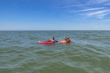 Bronzlaşmış adam denizde pembe şişme yatakta dinleniyor. Güneş gözlüklü başarılı adam tatilin ve serin içkilerin tadını çıkarıyor.